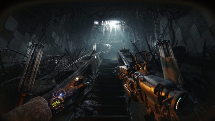 First-person view through a ruined subway tunnel with damaged escalator, barbed wire, and a futuristic rifle in the foreground, dim light ahead.