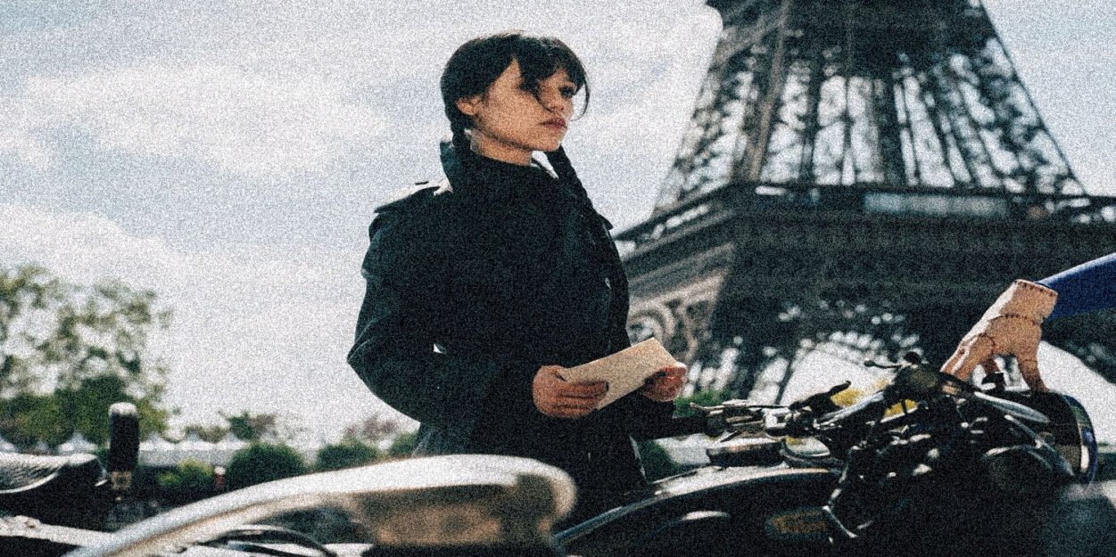 Woman in a black coat stands beside a bicycle, holding a note with the Eiffel Tower visible in the background.