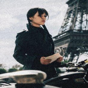 Woman in a black coat stands beside a bicycle, holding a note with the Eiffel Tower visible in the background.