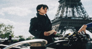 Woman in a black coat stands beside a bicycle, holding a note with the Eiffel Tower visible in the background.