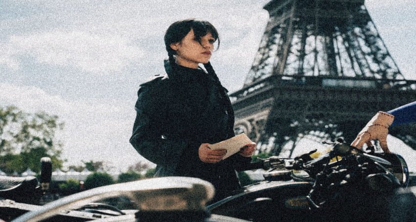 Woman in a black coat stands beside a bicycle, holding a note with the Eiffel Tower visible in the background.