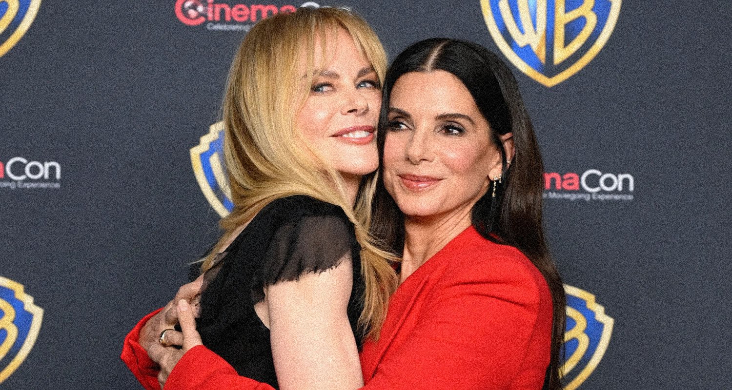 Two women hug on a red carpet at CinemaCon, with Warner Bros. logos in the background and one wearing a red outfit.