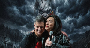 Two people posing on stage; a woman wearing a crown holds a microphone while embracing a man, both smiling warmly against a dark stormy backdrop.