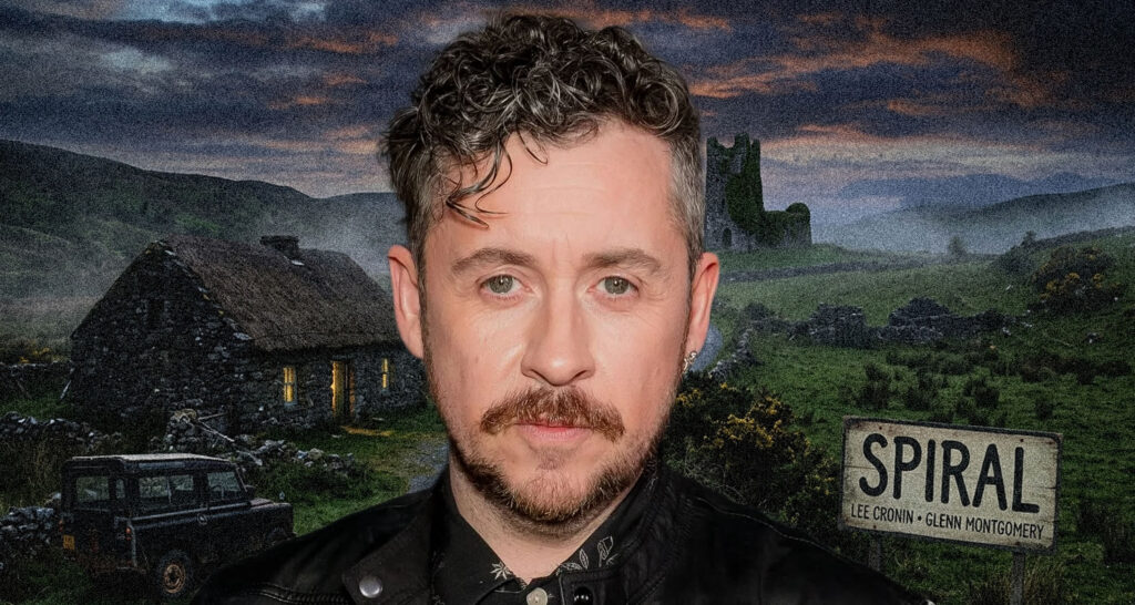 Portrait of a man with short dark hair, a mustache and beard, wearing a black jacket, against a rural backdrop with a sign that reads 'SPIRAL' behind him