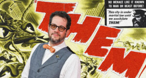 Portrait of a man with glasses and a beard, wearing a gray checked vest and orange bow tie, posed in front of a yellow collage with large red letters in the background.