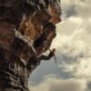 Climber hanging from a steep rock face against a cloudy sky, gripping with one arm.