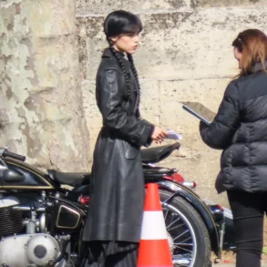 Two women stand by a parked motorcycle on a street; one in a long black coat with braided hair, the other holds a clipboard and talks to her.