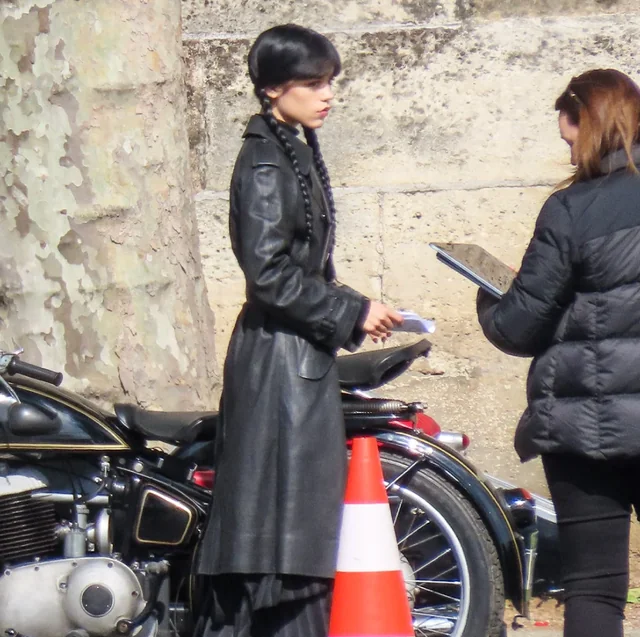Two women stand by a parked motorcycle on a street; one in a long black coat with braided hair, the other holds a clipboard and talks to her.