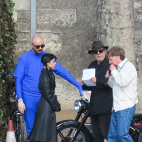 Group of four people beside classic motorcycles against a stone wall; a man in blue overalls points toward a woman in a black coat as two men review a sheet.