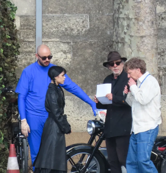 Group of four people beside classic motorcycles against a stone wall; a man in blue overalls points toward a woman in a black coat as two men review a sheet.