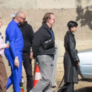 People walking in a line outdoors beside a concrete wall, some in casual clothes and one in a blue jumpsuit road-side scene