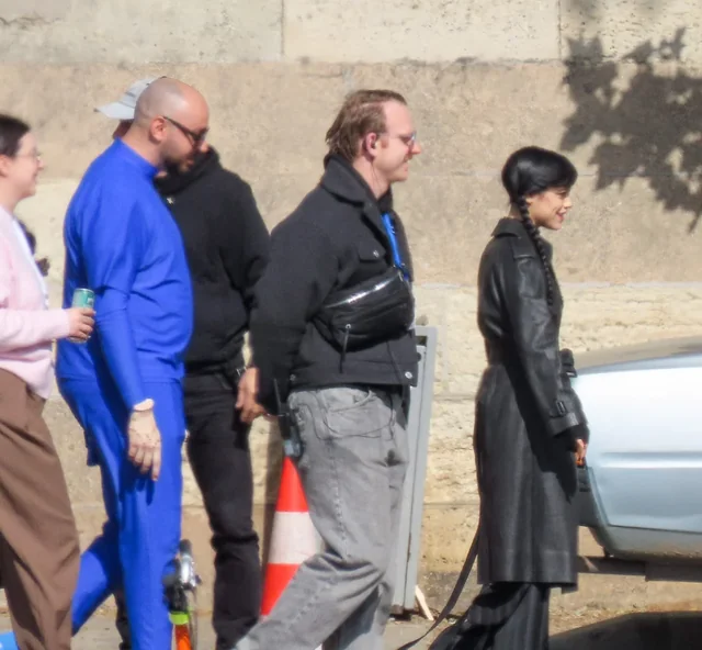 People walking in a line outdoors beside a concrete wall, some in casual clothes and one in a blue jumpsuit road-side scene