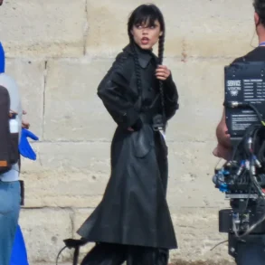 Woman in a long black trench coat with two braided pigtails stands on a film set beside camera gear against a stone wall.