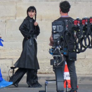 A film crew scene: woman in a black outfit with braided hair stands near a large camera rig outdoors against a stone wall: she looks to the side.