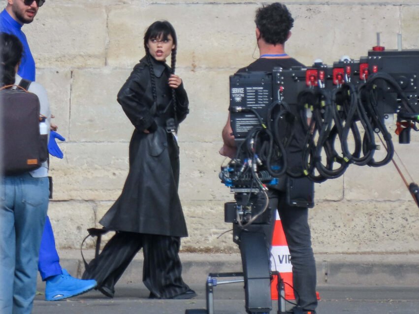 A film crew scene: woman in a black outfit with braided hair stands near a large camera rig outdoors against a stone wall: she looks to the side.