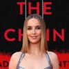 Portrait of a woman on the red carpet, standing in front of a black-and-red poster reading 'THE CHAIN' with hanging chains imagery.