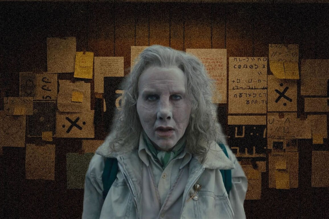 Woman with pale makeup and gray hair in a light jacket stands in a dim room with yellow sticky notes on a wall behind.
