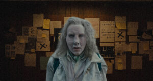 Woman with pale makeup and gray hair in a light jacket stands in a dim room with yellow sticky notes on a wall behind.