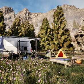Camping trailer with blue tarp, parked near wildflowers and pine trees against rocky mountains backdrop.