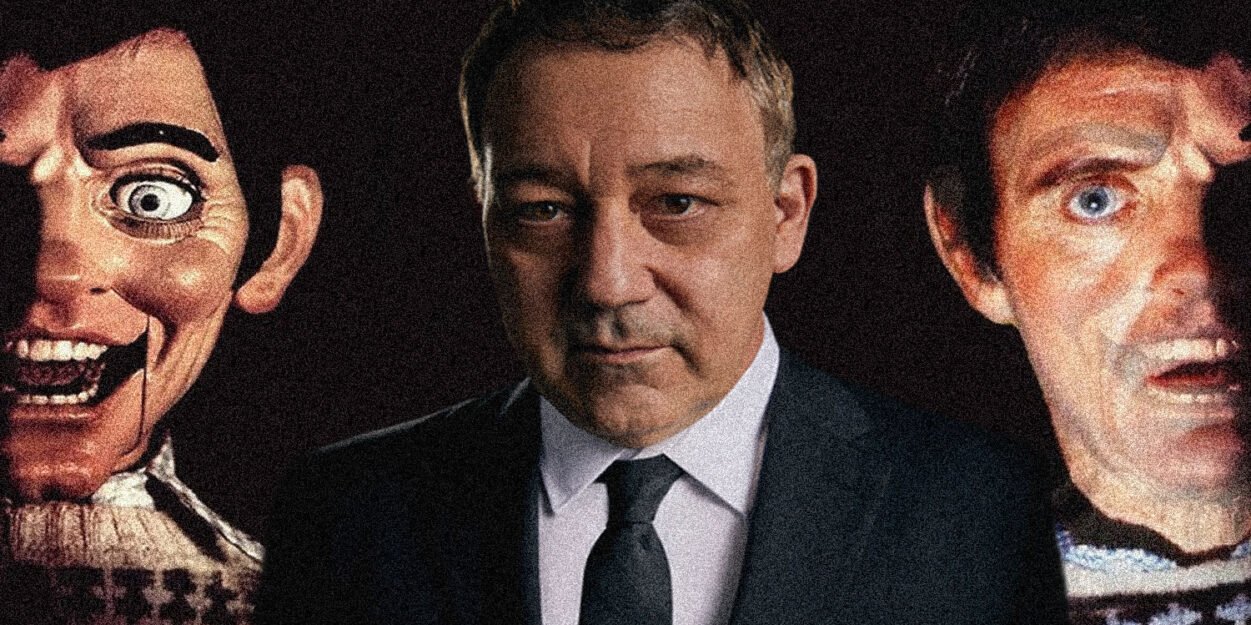 A man in a suit stands between two eerie, oversized mask faces in a dark setting, creating a unsettling portrait