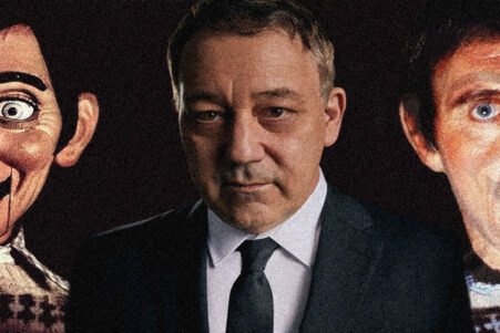 A man in a suit stands between two eerie, oversized mask faces in a dark setting, creating a unsettling portrait