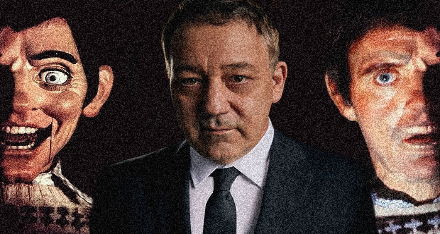 A man in a suit stands between two eerie, oversized mask faces in a dark setting, creating a unsettling portrait
