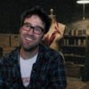 Smiling man with glasses in a plaid shirt sits in a dim workshop beside a dress form and shelves of tools and materials.