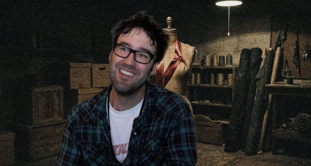 Smiling man with glasses in a plaid shirt sits in a dim workshop beside a dress form and shelves of tools and materials.