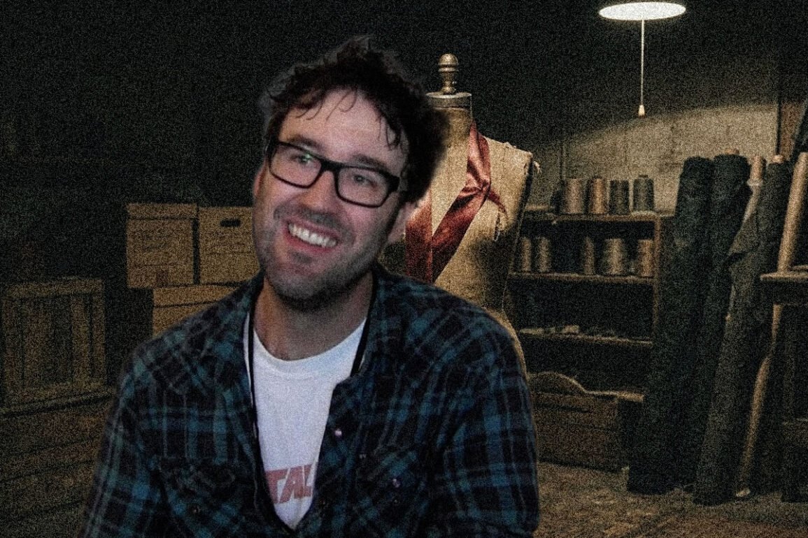Smiling man with glasses in a plaid shirt sits in a dim workshop beside a dress form and shelves of tools and materials.