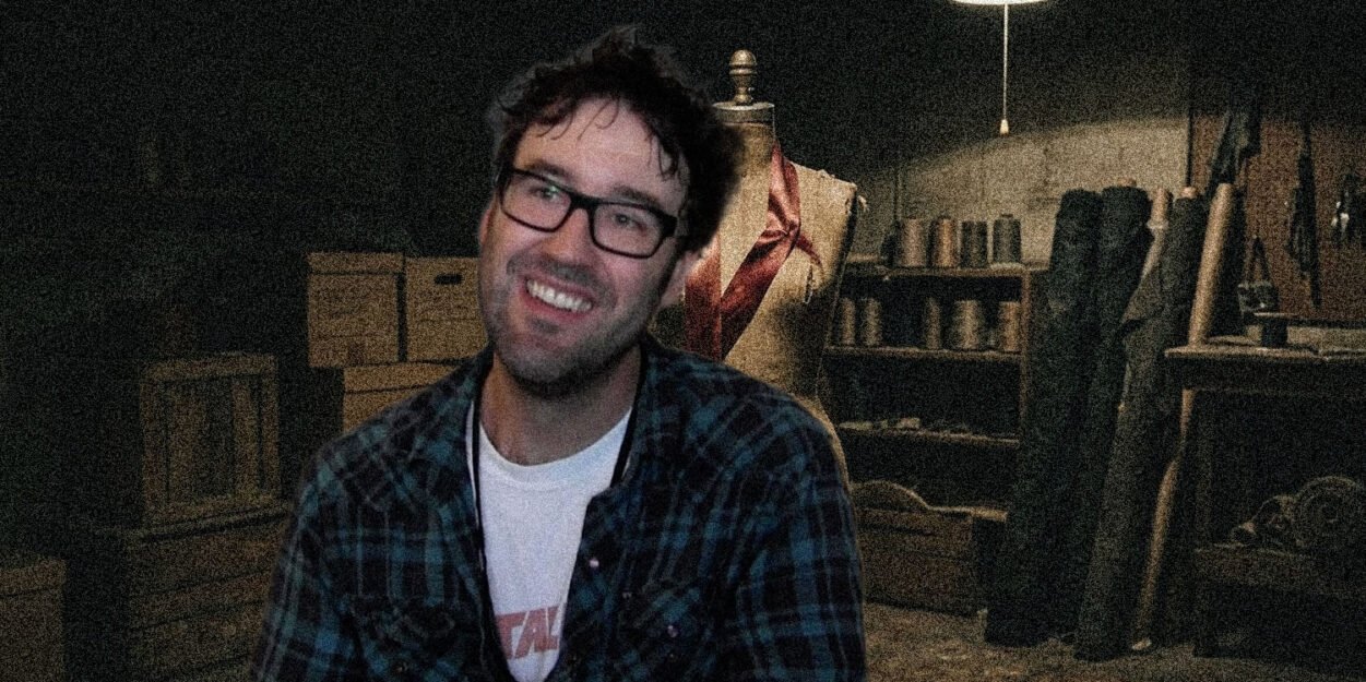 Smiling man with glasses in a plaid shirt sits in a dim workshop beside a dress form and shelves of tools and materials.