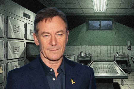 Man in a dark blazer with a yellow ribbon lapel pin stands in a vault-like room filled with metal safes and a large table in the background.