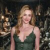 Woman in a green lace dress posing in a dim, cluttered workshop filled with mannequin torsos and heads under hanging bulbs.