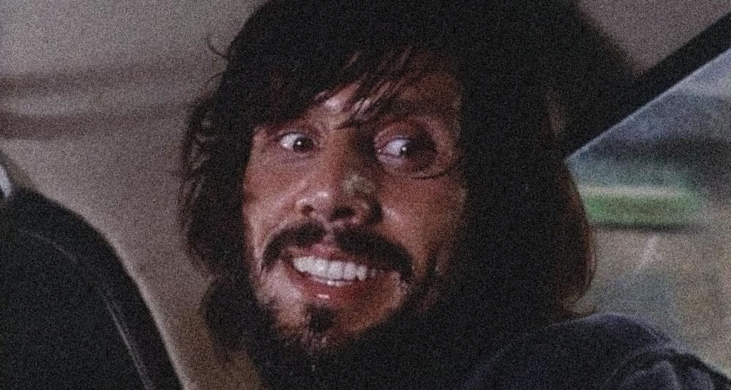 Man with dark hair and a beard smiling while seated in a car at night, looking to his left.