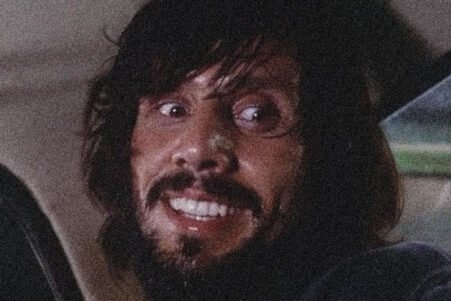 Man with dark hair and a beard smiling while seated in a car at night, looking to his left.