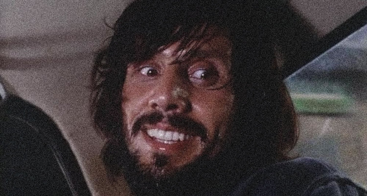Man with dark hair and a beard smiling while seated in a car at night, looking to his left.