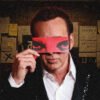 Man in a suit holds a red blindfold over his eyes, standing in a dim room covered with sticky notes and papers behind him.