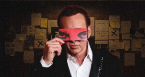 Man in a suit holds a red blindfold over his eyes, standing in a dim room covered with sticky notes and papers behind him.
