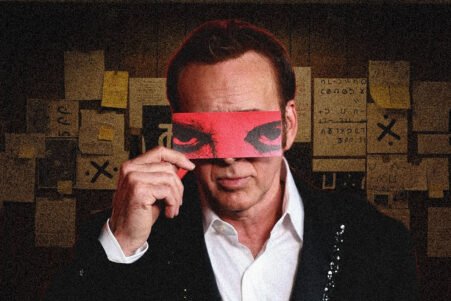Man in a suit holds a red blindfold over his eyes, standing in a dim room covered with sticky notes and papers behind him.