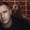 Moody portrait: man in foreground with pale skin, blue eyes, dark shirt; a horned demon figure looms behind him in a dim, grid-patterned background.