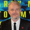 Portrait of a man in a dark suit and tie with a red lapel pin, against a blue smoke backdrop and large yellow text behind him.