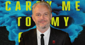 Portrait of a man in a dark suit and tie with a red lapel pin, against a blue smoke backdrop and large yellow text behind him.