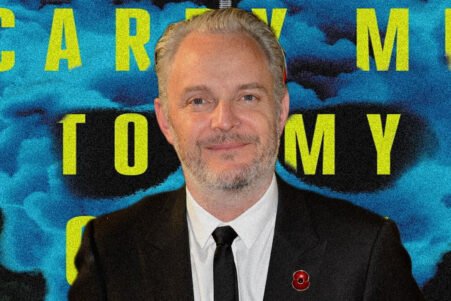 Portrait of a man in a dark suit and tie with a red lapel pin, against a blue smoke backdrop and large yellow text behind him.