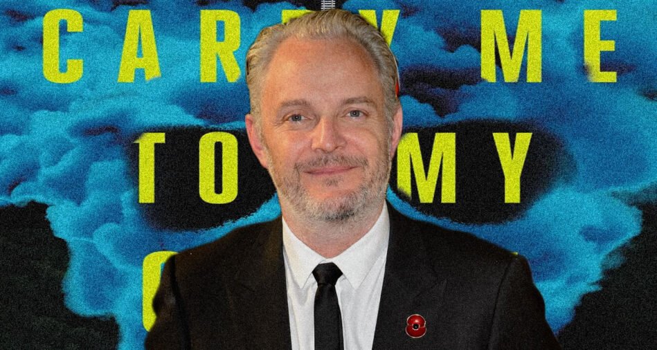 Portrait of a man in a dark suit and tie with a red lapel pin, against a blue smoke backdrop and large yellow text behind him.
