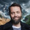 Smiling man with a beard in front of a surreal collage of desert dunes, snow-capped mountains, and a tropical jungle.