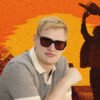 Man in sunglasses posed in front of an orange sunset collage with a windmill and a silhouette raising a trophy in the background.
