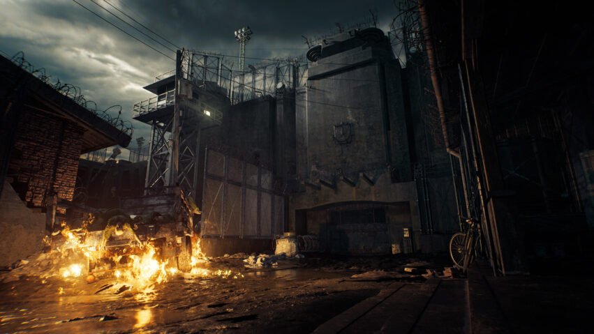 Ominous detention complex at dusk with tall concrete walls, barbed wire, and a fire burning in the rubble at the front.