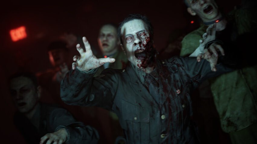 Zombie man in a blood-splattered shirt reaches toward the camera with outstretched hands in a dark, red-lit scene.