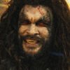 Zombie-like man with long hair, torn forehead wound, and yellowed fangs snarling in a smoky, dimly lit scene.