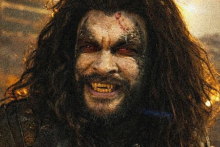 Zombie-like man with long hair, torn forehead wound, and yellowed fangs snarling in a smoky, dimly lit scene.