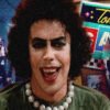 Smiling person wearing a green top and chunky pearl necklace, foreground of a neon arcade/game night scene with masked figures in the background.